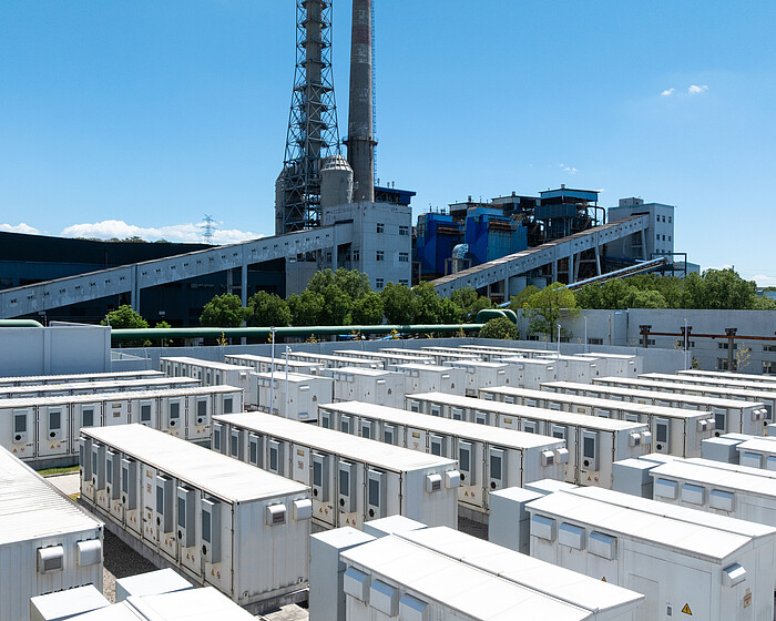 An aerial view of rows of white battery energy storage systems under a clear blue sky with a large industrial building in the background An aerial view of rows of white battery energy storage systems under a clear blue sky with a large industrial building in the background.